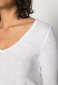 Balta V formos kaklo palaidinė, pagaminta iš lengvos, tekstūruotos medžiagos. Turi trumpas rankoves ir laisvą siluetą, atidengiantį odą prie raktikaulio.