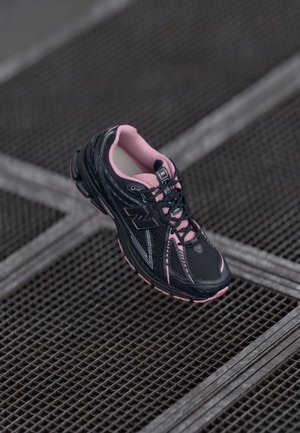Black and pink New Balance running shoe with laces on a metal grid surface, angled to show side and top details.