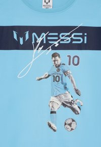 Gaiši zils T-krekls ar grafiku, kurā attēlots futbolists akcijā, ar vārdu "MESSI" un numuru "10" tumši zilā krāsā.