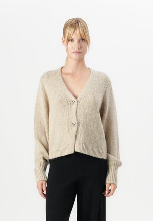 Blond kvinna som bär en beige stickad kofta med två dekorativa spännen och svarta byxor, står mot en plain vit bakgrund.