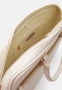 Beige taske med en glat overflade og lynlåslukning, der har en indvendig lomme og et logo-tag. Robuste håndtag i en matchende farve.