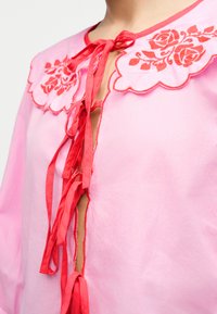 Ροζ μπλούζα με μεγάλο γιακά με κυματιστό τελείωμα που διαθέτει κεντήματα με κόκκινα τριαντάφυλλα και πολλαπλές κόκκινες κορδέλες δεμένες κατά μήκος της μπροστινής πλευράς.