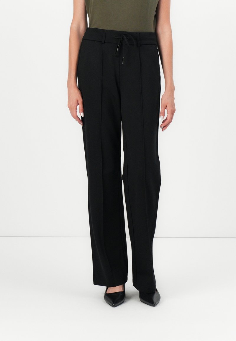 Femme portant un pantalon large noir sur mesure avec des coutures devant, une taille à cordon, un top vert sans manches et des chaussures noires pointues.