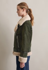 Groene suède jas met een zachte beige schapenvacht kraag en manchetten; beschikt over een voorkant met ritssluiting en een ceintuur in de taille voor extra detail.