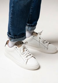 Witte leren sneakers met een textuurafwerking, voorzien van een bruine hakaccent, witte veters en een minimalistisch ontwerp. G gedragen met opgerolde denim.