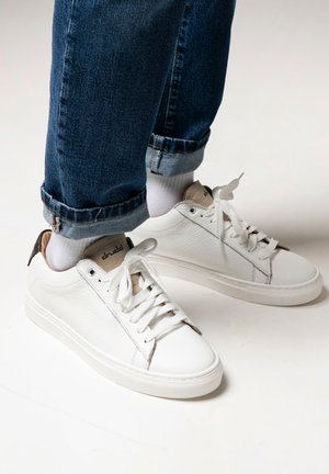 Sneakers bianchi in pelle con una finitura testurizzata, caratterizzati da un accento di tacco marrone, lacci bianchi e un design minimalista. Indossati con jeans risvoltati.