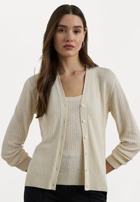 Cardigã canelado beje claro com decote em V, frente botonada e mangas longas. Textura brilhante e tecido suave. Usado sobre uma blusa correspondente.