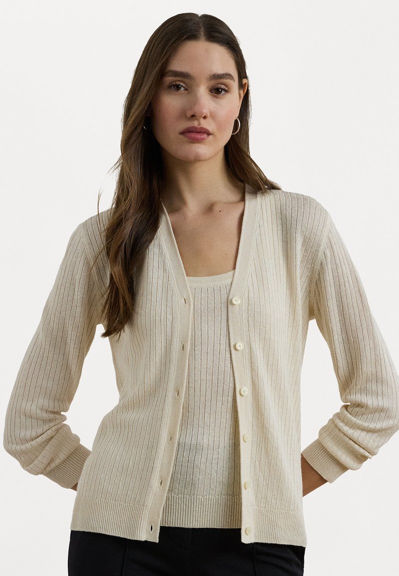 Cardigã canelado beje claro com decote em V, frente botonada e mangas longas. Textura brilhante e tecido suave. Usado sobre uma blusa correspondente.