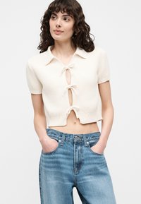 Jeune femme aux cheveux bouclés portant un cardigan court crème à manches courtes avec trois liens devant et un jean bleu taille haute.