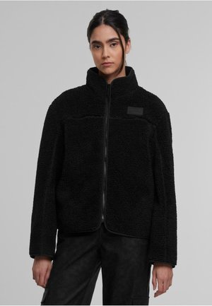 Giacca in pile nera con colletto alto, chiusura con zip e superficie testurizzata. Presenta una piccola toppa con logo sul petto, vestibilità rilassata.