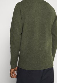 Person som bär en långärmad, olivgrön stickad tröja med svarta byxor, sedd bakifrån mot en enkel ljus bakgrund.