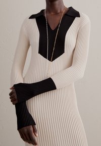 Abito in maglia a coste con una combinazione di colori nero e crema, dotato di scollo a V, colletto e maniche allungate con aperture per le dita.
