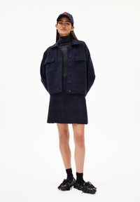 Marineblauwe wolmixjas met knopenfront en twee paspelzakken, gecombineerd met een bijpassende geruite rok en schoenen met zwarte franjes.
