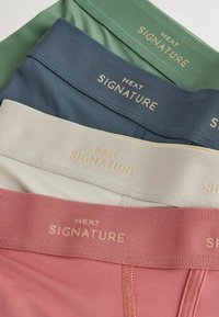 Četiri para donjeg rublja u bojama zelene, mornarske, bež i koraljne. Svaki par ima mekani, elastični pojas s natpisom "NEXT SIGNATURE" u zlatnim slovima.