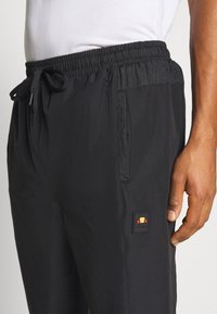Zwarte broek met elastische tailleband, gemaakt van lichtgewicht stof; beschikt over zijzakken en een klein merklabel met een geel en rood ontwerp.
