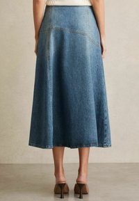 Lichteblauwe denim midi-rok met een uitlopende vorm, voorzien van dubbele stiksels en een onafgewerkte zoom. Draagt zich met beige hakken.