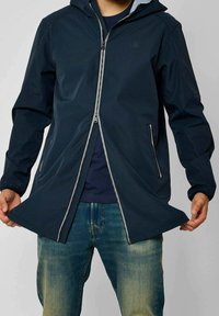 Námořnická nepromokavá bunda s kapucí, vybavená předním zipem, dvěma bočními kapsami a elastickými manžetami, v kombinaci s modrými džínami.