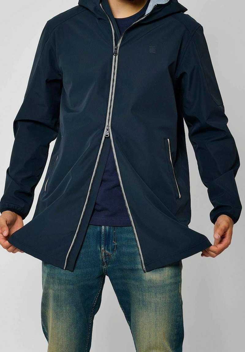 Námořnická nepromokavá bunda s kapucí, vybavená předním zipem, dvěma bočními kapsami a elastickými manžetami, v kombinaci s modrými džínami.