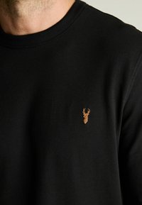 Sudadera de algodón negra con cuello redondo, que presenta un pequeño diseño de ciervo bordado en marrón en la zona del lado izquierdo del pecho.