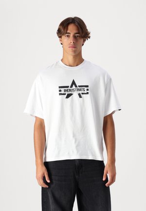 Young man with brown hair wears white oversized T-shirt with black "INDUSTRIES" logo and black pants, standing against plain background.