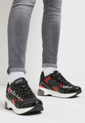 Personne portant des baskets Replay noires et rouges avec semelles blanches, associées à des chaussettes blanches et un jean gris retroussé, debout sur un sol clair.