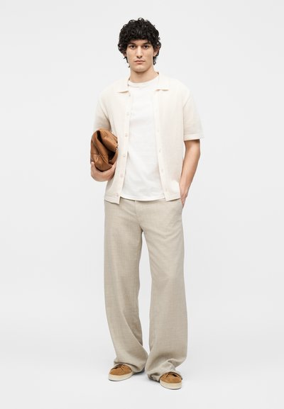 Νέος άνδρας με σγουρά μαλλιά, που φοράει εκρού πουκάμισο και μπεζ φαρδιά παντελόνια, κρατώντας μια διπλωμένη καφέ δερμάτινη τσάντα σε λευκό φόντο.