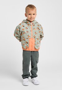 Veste légère à capuche vert menthe avec des accents orange et un motif de limace de dessin animé ; associée à un pantalon gris et des baskets blanches.
