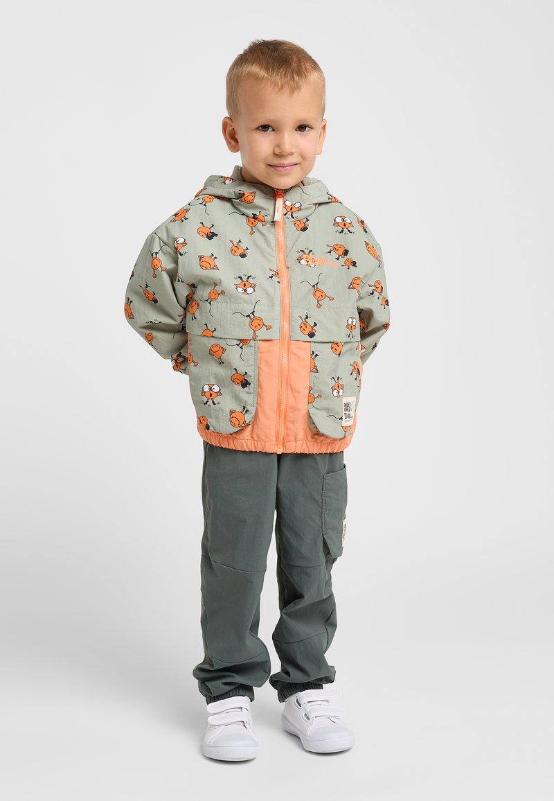 Veste légère à capuche vert menthe avec des accents orange et un motif de limace de dessin animé ; associée à un pantalon gris et des baskets blanches.