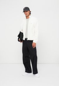 White fleece jacket, gray t-shirt, black cargo pants, and white sneakers; wearing a dark baseball cap with a logo. Holding a black jacket.