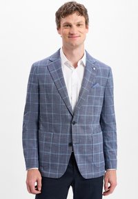Man in een blauwe geruite blazer, witte overhemd en donkere broek, glimlachend tegen een witte achtergrond.