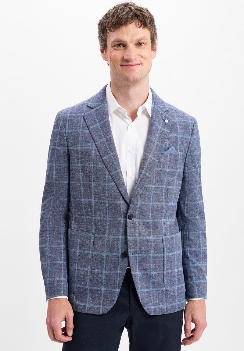 Man in een blauwe geruite blazer, witte overhemd en donkere broek, glimlachend tegen een witte achtergrond.