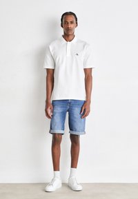 Vit polo skjorta med krage, denim shorts rullade vid fållen och vita sneakers. Modellen står framför en neutral bakgrund.