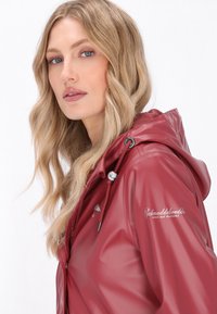 Burgundy vettita säänkestävä takki, jossa on huppu, joustava kiristin ja logo-yksityiskohta hihassa, sekä kiiltävä ja sileä pinta.