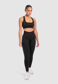 Sort sports-bh og leggings med høj talje lavet af elastisk stof. Har en glat tekstur og en tætsiddende silhuet. Bæres med hvide sneakers.