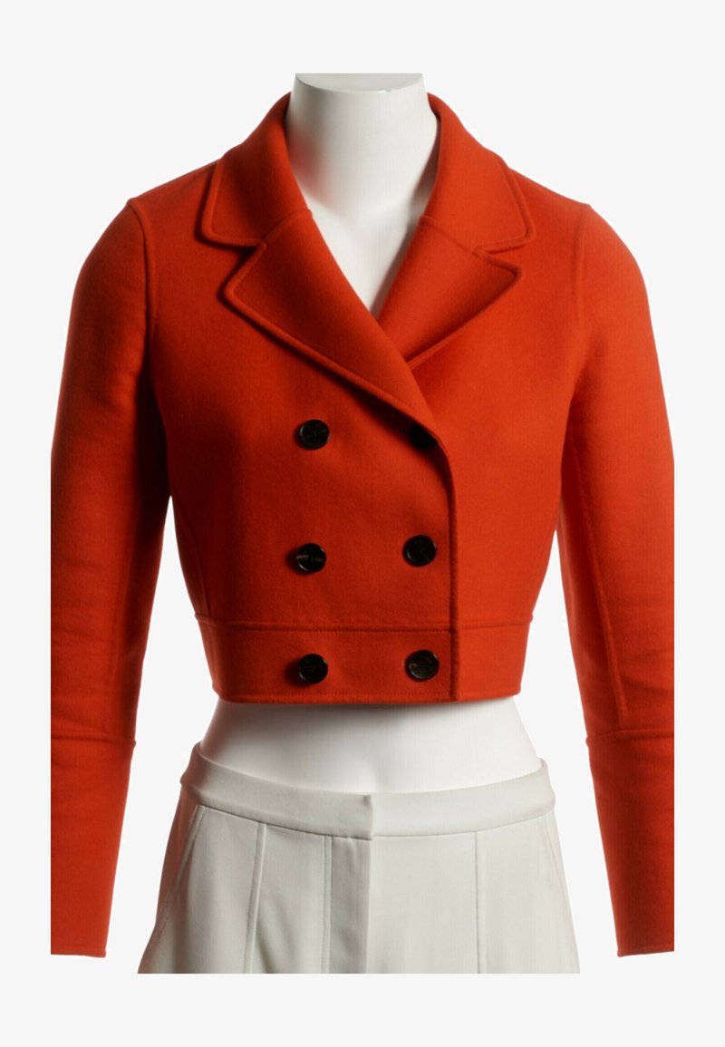 Veste orange courte croisée à doubles boutons noirs associée à un pantalon blanc plissé taille haute sur un mannequin.