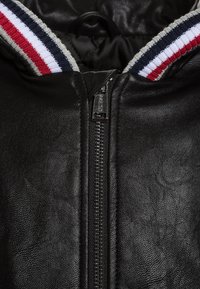 Nærbillede af sort textured læderjakke, der viser lynlåsen med "Guess" branding og ribbet krave med røde, hvide og marineblå striber.