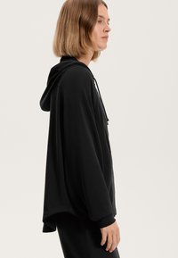 Črna kapuca s sproščenim oblikovanjem in mehkimi vlakni. Ima vrvico in dolge rokave, ki segajo pod pas.