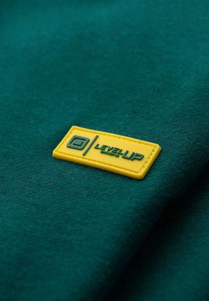 Tela verde con una textura suave que presenta un parche rectangular de goma amarilla con la inscripción "LEVEL UP" y un diseño de logo.