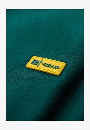 Vihreä kangas, jossa on sileä pinta, sisältää suorakaiteen muotoisen, keltaisen kumitarran, jossa on kohokuvioitu teksti "LEVEL UP" ja logo.