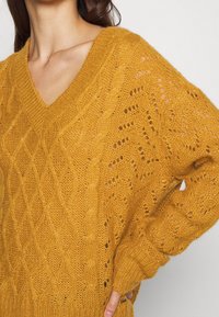 Mosterdgele gebreide trui met een V-hals, voorzien van een kabelpatroon en opengewerkte details. Wijde mouwen met ribgebreide manchetten.