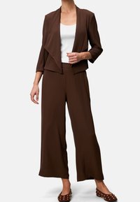 Ruskeat leveälahkeiset housut, jotka on yhdistetty vastaavaan laskeutuvaan bleiseriin valkoisen topin kanssa. Asussa on pehmeä kangas ja rento istuvuus.