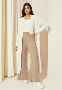 Cardigan beige chiaro con maniche ampie e tasche frontali, abbinato a pantaloni larghi color cammello e sneakers bianche. Texture liscia; design minimal.