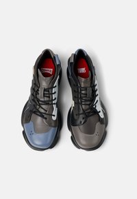 Due scarpe da ginnastica realizzate con materiali misti. La scarpa sinistra è blu e grigia, la destra è nera e marrone. Entrambe presentano lacci neri e accenti rossi all'interno.