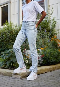 Bílá tričko s jemným logem, světle modré džíny s vysokým pasem a robustní bílé tenisky s růžovými akcenty, zasazené do zeleně a květin.