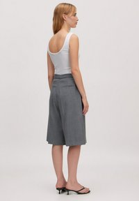 Vrouw staat zijwaarts gekeerd in een witte mouwloze top, grijze, knielange wijde shorts en zwarte hoge hakken tegen een effen achtergrond.