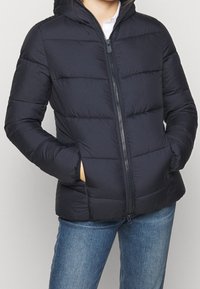 Veste matelassée en bleu marine, avec fermeture éclair à l'avant, capuche et poches latérales. Le tissu a une texture lisse avec des rayures horizontales matelassées.