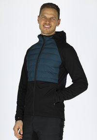 Letvægts hættejakke med en teal polstret overdel og sort strækstof. Har fuld lynlås, sidelommer og en tætsiddende design.