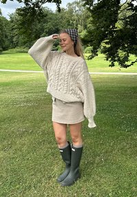 Cremefarbener Kabelstrickpullover mit übergroßen Ärmeln, beiger Minirock und grünen Gummistiefeln. Haare mit einem schwarz-weißen karierter Haarband zusammengebunden.