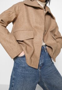 Tan uldblandet jakke med krave, to forlommer og knaplukning, parret med højtaljede blå denim jeans.