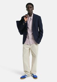 Blazer en tissu marine, chemise à rayures légères, pantalon crème et baskets bleues avec accents blancs. Modèle tenant un sac fourre-tout marine.